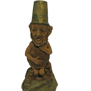 May include: A small, brown ceramic gnome figurine wearing a green and gold thimble hat. The gnome is holding a brown ball in its hands and is standing on a green base with the words "THREADBARE" and "1997" inscribed on it.
