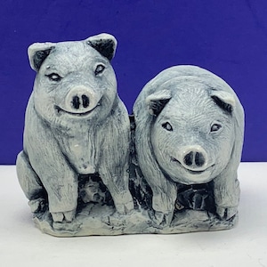 May include: Two gray ceramic pig figurines sitting side by side on a white base. The pigs have a smiling expression and are looking at the viewer.