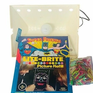 May include: A Lite-Brite Picture Refill box featuring Super Friends characters. The box includes a clear plastic tray with a light bulb, a bag of colorful pegs, and a picture template featuring Superman, Wonder Woman, and Batman. The box is labeled "Lite-Brite Picture Refill" and "Super Friends".