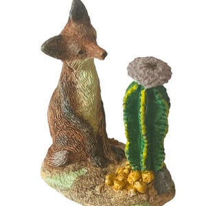 Puede incluir: Una figurita de zorro marrón y blanco con una figurita de cactus verde y amarillo. El zorro está mirando al cactus. Las figuritas están sobre una base marrón.
