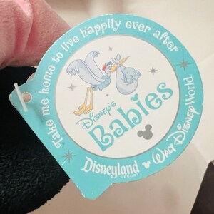 May include: A blue and white tag with the text "Take me home to live happily ever after Disney's Babies Disneyland Walt Disney World Resort" and a cartoon image of a stork carrying a baby.