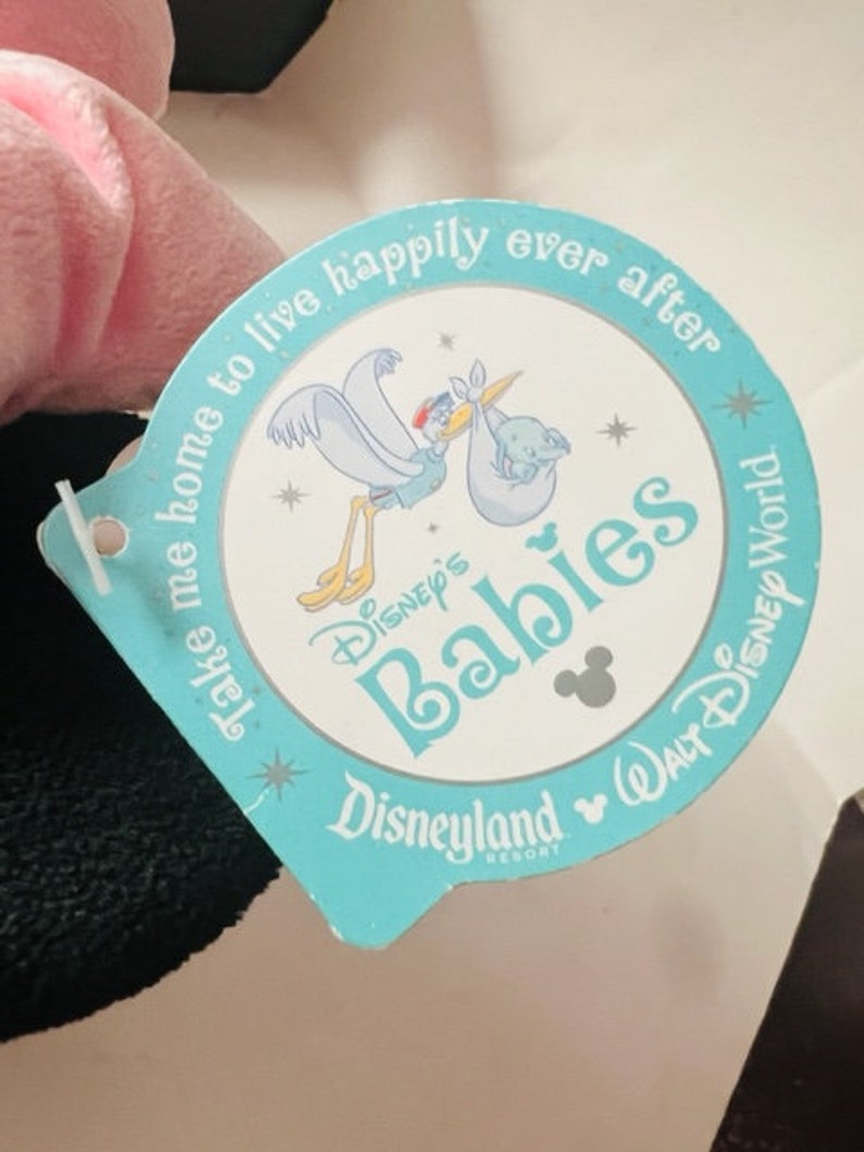May include: A blue and white tag with the text "Take me home to live happily ever after Disney's Babies Disneyland Walt Disney World Resort" and a cartoon image of a stork carrying a baby.