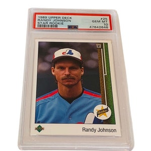 Puede incluir: Una tarjeta de béisbol Randy Johnson Star Rookie de 1989 Upper Deck, calificada GEM MT 10, en un soporte protector. La tarjeta presenta a Randy Johnson con un uniforme de béisbol azul y una gorra blanca.