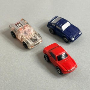 May include: Three miniature toy cars including one clear plastic vehicle with visible internal mechanical details, one solid dark blue car with a silver roof panel, and one solid red car, all featuring black wheels and moulded body designs.