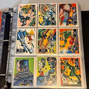 Könnte beinhalten: Eine Sammlung von Batman-Sammelkarten im Vintage-Stil in einem Ordner. Die Karten zeigen farbenfrohe Illustrationen von Batman und Robin in Aktion, mit Sprechblasen und Aktionswörtern wie "BAM" und "BOOM". Die Karten befinden sich in Schutzhüllen.