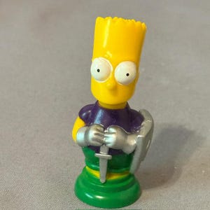 May include: A yellow Bart Simpson figurine in a knight's costume, holding a silver sword and shield. The figurine has a green base and a purple tunic. The figure is a collectible item.