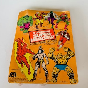 May include: A yellow card with images of various superheroes, including The Thing, The Human Torch, Conan, and Falcon. The card reads "Les Extraordinaires Super-Heroes! Collectionne Les Tous".