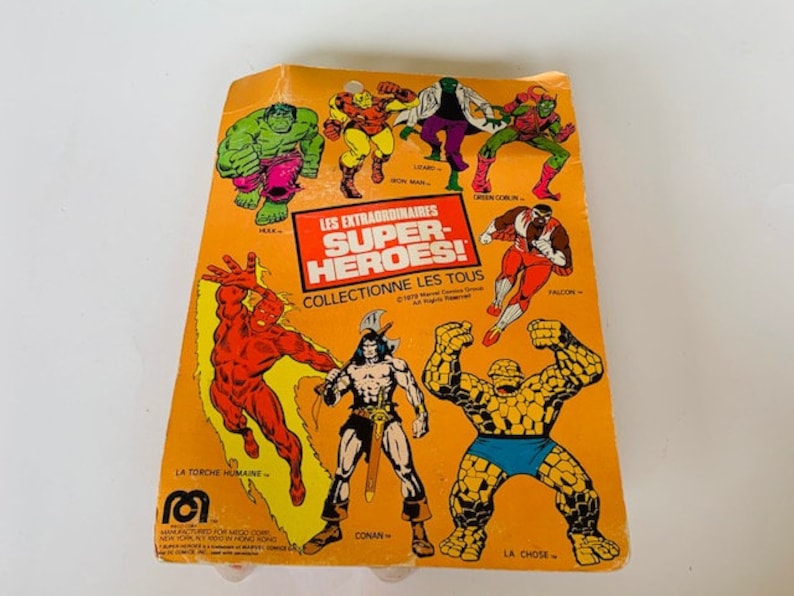 May include: A yellow card with images of various superheroes, including The Thing, The Human Torch, Conan, and Falcon. The card reads "Les Extraordinaires Super-Heroes! Collectionne Les Tous".