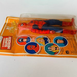 May include: A vintage action figure of Spider-Man in his red and blue suit, packaged in a plastic blister pack. The packaging features a yellow and orange background with images of other superheroes, including Captain America, Thor, and Spider-Man. The text on the packaging reads "Les Extraordinaires Super Heroes! Spider-Man".