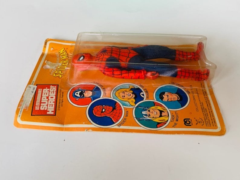 May include: A vintage action figure of Spider-Man in his red and blue suit, packaged in a plastic blister pack. The packaging features a yellow and orange background with images of other superheroes, including Captain America, Thor, and Spider-Man. The text on the packaging reads "Les Extraordinaires Super Heroes! Spider-Man".