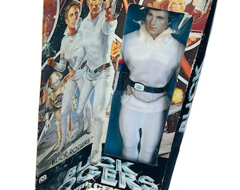 May include: A vintage Buck Rogers in the 25th Century action figure in a white jumpsuit with a black belt and a silver gun. The figure is in a cardboard box with a colorful image of Buck Rogers and other characters.