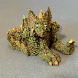 Peut inclure: Figurine détaillée d'un dragon vert et beige aux yeux bleus. Le dragon est allongé, les bras et les jambes tendus. La figurine a un aspect texturé et écailleux, sur fond gris.