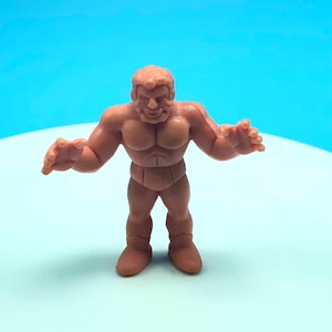 May include: A brown rubber figurine of a muscular man with a short haircut. The figurine is posed with arms outstretched and a determined expression.