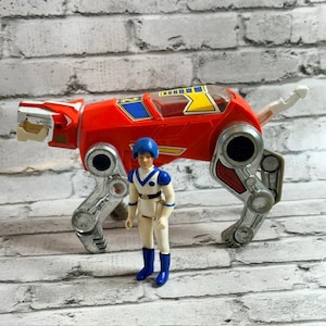 May include: A red and silver robotic lion toy featuring blue and yellow accents on its back, accompanied by a small action figure wearing a white suit with blue boots, gloves, and a blue helmet, standing in front of the mechanical animal figure.