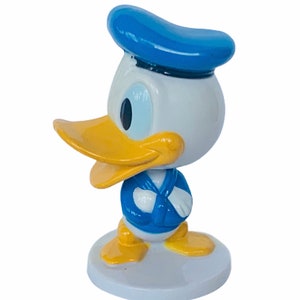 May include: A small, blue and yellow figurine of Donald Duck wearing a blue sailor hat. The figurine is standing on a white base.