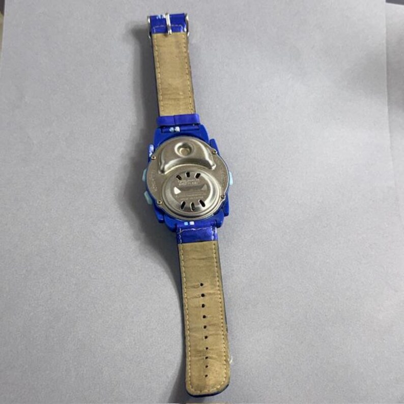 Puede incluir: Un reloj digital azul con una correa de cuero marr&oacute;n claro. La parte trasera del reloj es plateada. La correa es de color marr&oacute;n claro con una serie de agujeros para el ajuste.