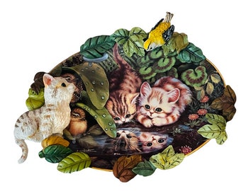 May include: A ceramic wall plaque featuring a whimsical scene of three kittens and two birds nestled in a lush green garden. The kittens are depicted in a playful pose, with one looking up at a bird perched on a branch. The plaque is decorated with intricate details, including leaves, flowers, and a small stream.