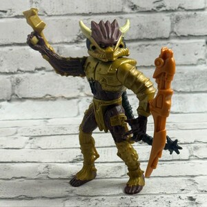 Peut inclure: Une figurine en plastique représentant une créature au corps blindé marron et doré, dotée d'une tête à cornes, d'un bras droit levé tenant une petite arme dorée et d'un bras gauche tenant un grand accessoire orange en forme de bouclier.