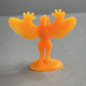 Peut inclure: Petite figurine en plastique orange représentant un oiseau aux ailes déployées. La figurine a une base ronde et un design stylisé, avec un corps et des ailes légèrement translucides. La figurine est de couleur orange unie.