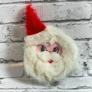 May include: A fuzzy Santa Claus face ornament featuring a bright red pointed hat, a thick white beard, and a moustache made of soft, textured material. The face has pink fabric skin with embroidered black eyes and a small red stitched mouth.