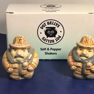 May include: Two ceramic salt and pepper shakers shaped like firefighters. The shakers are wearing brown and gray uniforms with yellow accents. The shakers are standing on a white surface. The box they came in is visible in the background. The box is white with black text that reads "Pot Bellys Salt & Pepper Shakers".