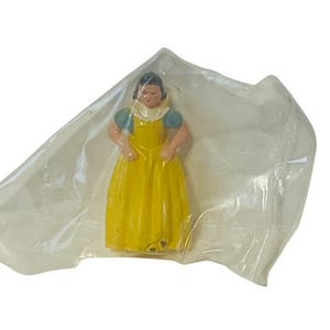 May include: A plastic figurine of Snow White, wearing a yellow dress with blue sleeves and a white collar. She is holding a small white object in her right hand.