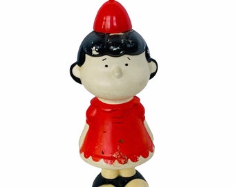 May include: A vintage plastic figurine of a girl with black hair wearing a red dress and a red hat. The figurine has a white face and is in good condition.