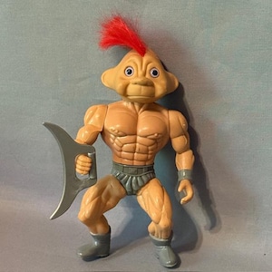 May include: A vintage action figure of a muscular humanoid alien with a red mohawk and a gray weapon. The figure is tan with gray shorts, wristbands, and boots. The toy has a detailed face and is posed in a fighting stance.