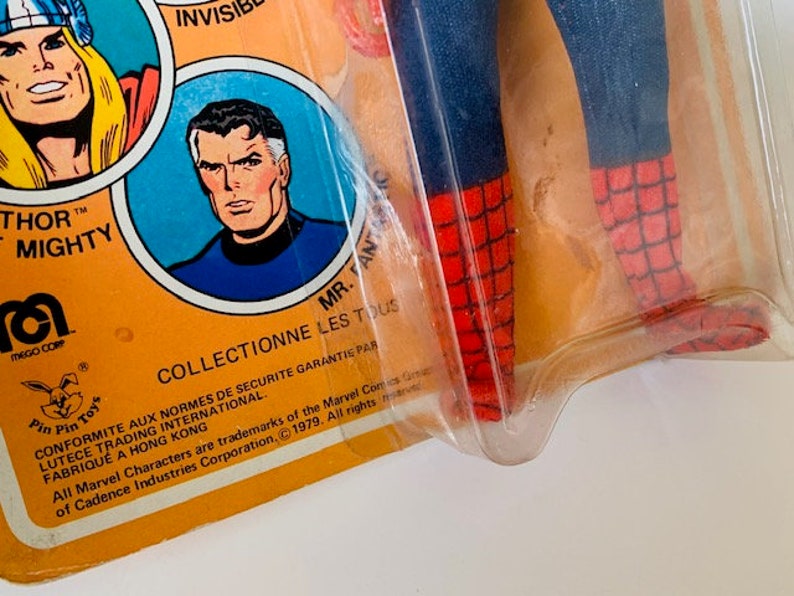 May include: A vintage action figure of Spider-Man in a red and blue suit with a web design. The figure is packaged in a plastic blister pack with a yellow cardboard backing. The backing features images of Thor and Mr. Fantastic, along with the Mego Corp logo and text that reads "Collectionne les tous" and "Conformite aux normes de securite garantie par Lutece Trading International. Fabrique a Hong Kong. All Marvel Characters are trademarks of the Marvel Comics Group, Inc. 1979. All rights reserved. of Cadence Industries Corporation."