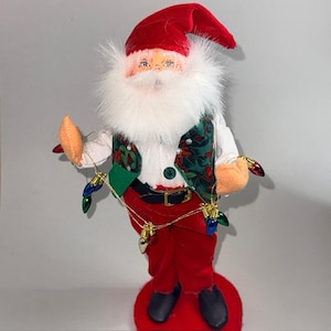 May include: A Santa Claus figurine with a red velvet hat and pants, a white beard, and a green vest with a floral pattern. The figurine holds a string of Christmas lights and has black shoes. The base is red.