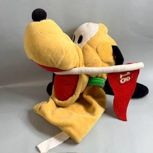 May include: A yellow plush dog character with a black nose, white eye patches, and a green collar. The character holds a white golf club shaft in its mouth, which is attached to a red triangular golf flag featuring the embroidered white number 18.