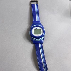 Puede incluir: Un reloj de pulsera digital azul con una correa azul a juego. La esfera del reloj es blanca con una pantalla digital. La correa tiene rayas blancas y azul claro. El reloj tiene una hebilla plateada.