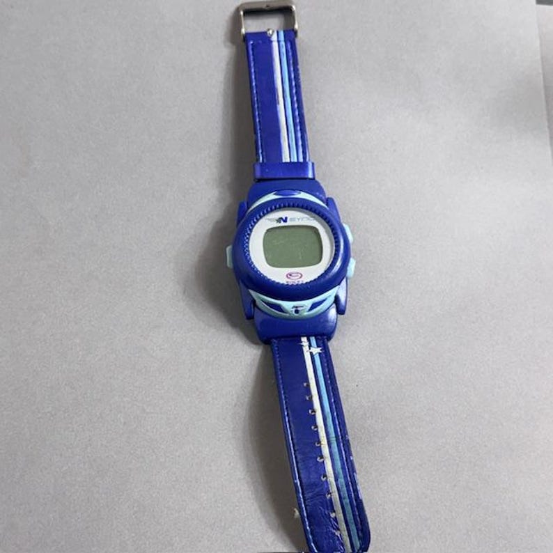 Puede incluir: Un reloj de pulsera digital azul con una correa azul a juego. La esfera del reloj es blanca con una pantalla digital. La correa tiene rayas blancas y azul claro. El reloj tiene una hebilla plateada.