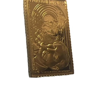May include: Gold-colored metal stamp with a raised image of two people, one holding a torch and the other holding a globe. The stamp has a serrated border and the text "UNITED STATES OF AMERICA" is visible at the top.