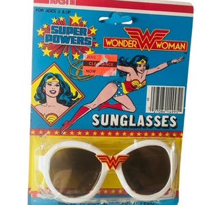 May include: Wonder Woman sunglasses in original packaging. The white-framed sunglasses have brown lenses and a red and gold emblem. The packaging is blue, red, and yellow, featuring Wonder Woman graphics and the text "SUPER POWERS" and "SUNGLASSES."