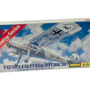 May include: A model airplane kit box featuring a white and gray Fieseler Fi 156 Storch aircraft with red and black markings. The box is labeled "Prestige Series" and "Aurora Heller".