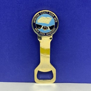 Op de afbeelding: Gouden flesopener met een blauw en gouden cirkelvormig ontwerp. Het ontwerp toont een handdruk boven een open boek met de tekst "Masons Don't Seek Men Men Seek Masonry" rond de rand.