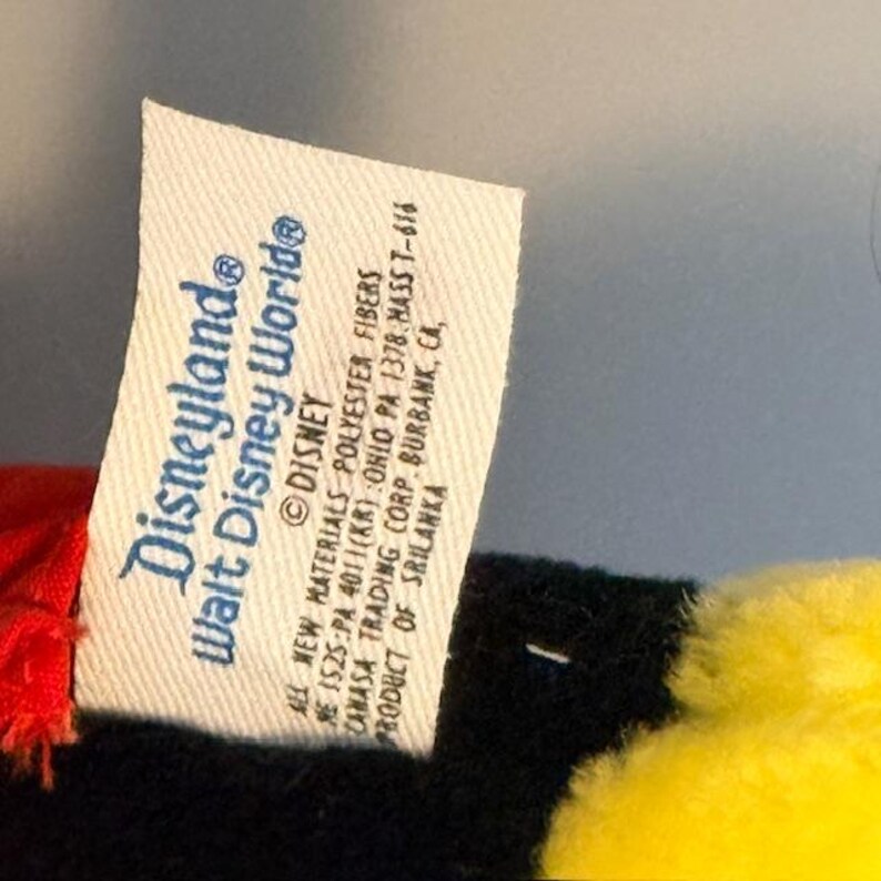 Pu&ograve; includere: Primo piano di un'etichetta bianca di un prodotto Disneyland Walt Disney World. L'etichetta presenta i loghi Disneyland e Walt Disney World in blu. L'etichetta include anche testo sui materiali e l'origine del prodotto.