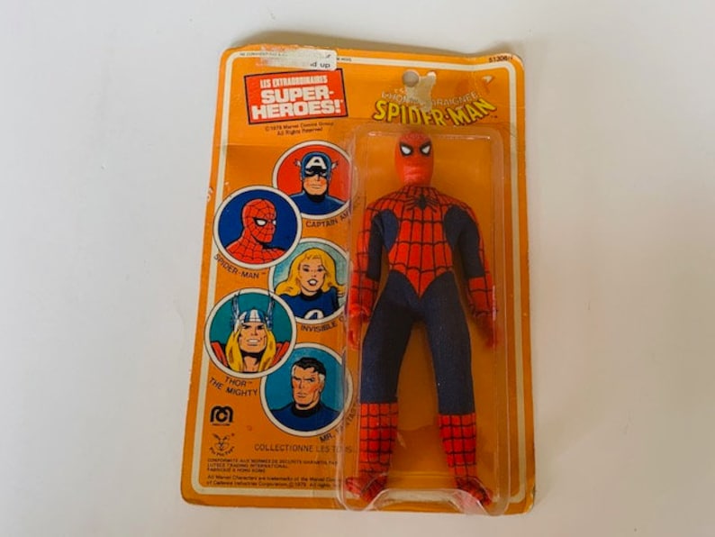 May include: A vintage action figure of Spider-Man in his red and blue suit with a spider web design. The figure is packaged in a plastic blister pack with a yellow and orange cardboard backing. The packaging features images of other superheroes, including Captain America, Spider-Man, Thor, and the Incredible Hulk. The text on the packaging reads "Les Extraordinaires Super-Heroes!" and "Spider-Man".