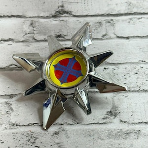 Pode incluir: Um emblema em forma de estrela com acabamento cromado e uma incrustação circular central. A incrustação apresenta um fundo amarelo brilhante com um design de cruz vermelha de quatro pontas delineada em azul. A estrela tem oito pontas.
