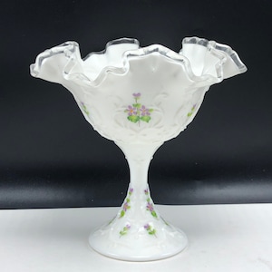 May include: A white milk glass candy dish with a ruffled edge and a silver trim. The dish is decorated with hand-painted purple flowers and green leaves.