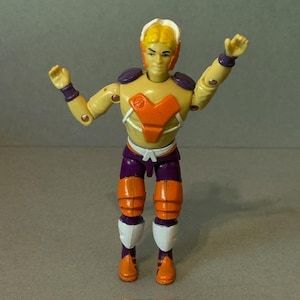 May include: A vintage action figure with a yellow and orange color scheme. The figure has a helmet, shoulder pads, and knee pads. The figure's arms are raised, and it is standing on a gray surface.