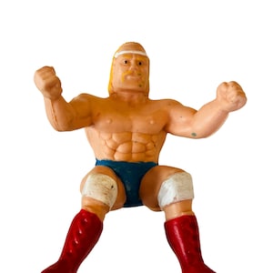 May include: A plastic action figure of a wrestler wearing a blue wrestling outfit with white knee pads and red boots. The figure has a yellow headband and a blonde wig.
