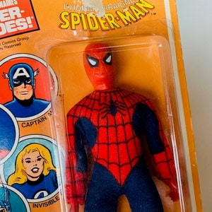 May include: A vintage action figure of Spider-Man in his red and blue suit with a black spider web design. The figure is packaged in a plastic blister pack with a yellow and orange cardboard backing. The packaging features the text "L'HOMME ARAIGNEE SPIDER-MAN" and "SUPER-HEROES!"