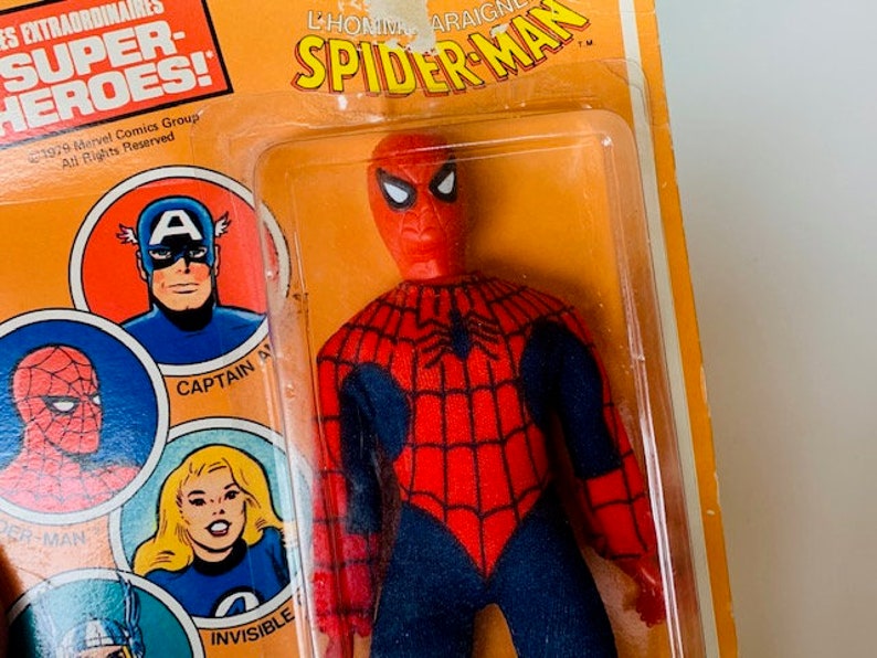 May include: A vintage action figure of Spider-Man in his red and blue suit with a black spider web design. The figure is packaged in a plastic blister pack with a yellow and orange cardboard backing. The packaging features the text "L'HOMME ARAIGNEE SPIDER-MAN" and "SUPER-HEROES!"