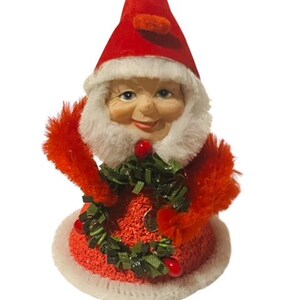 May include: A vintage Christmas elf ornament with a red hat, white beard, and a red and white dress with a green wreath.