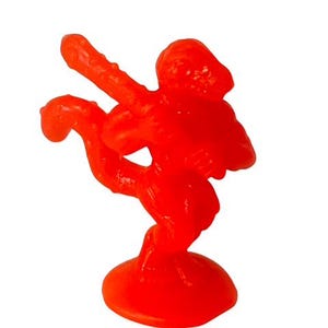 Peut inclure: Une figurine en plastique orange vif d'une personne en position de combat. La figurine est debout sur une base ronde.