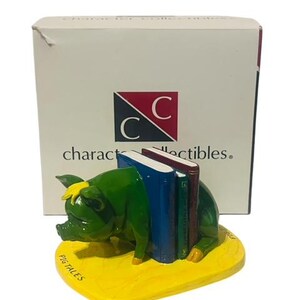 May include: Green pig bookends holding a stack of colorful books. The bookends are on a yellow base with the words "Pig Tales" written on it. The bookends are part of a collection called "Character Collectibles".
