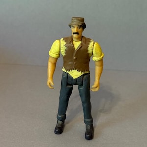 May include: A small, vintage action figure of a man wearing a brown hat, yellow shirt, brown vest, and dark blue pants. The figure has a mustache and black boots. The figure is standing upright.