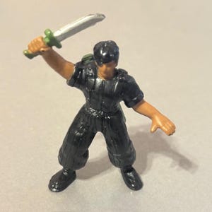 Pode incluir: Miniatura de uma figura de ação de uma pessoa em uniforme preto, segurando uma espada prateada e verde. A figura tem pele bronzeada e cabelo preto. A figura está em uma pose dinâmica, como se estivesse no meio de uma luta de espadas.